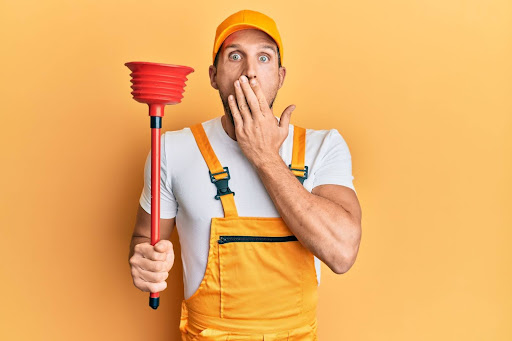 Homeowner dressed in orange overalls holding red plunger and gasping
